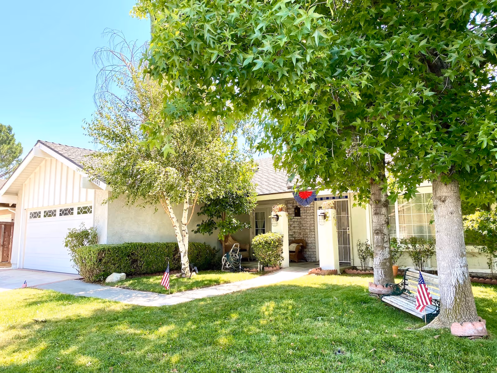 Front exterior view of a single-story house with a well-maintained lawn, two large trees, a bench with an American flag, and a driveway leading to a garage. The entrance has a small porch area with some outdoor seating and decorative plants.