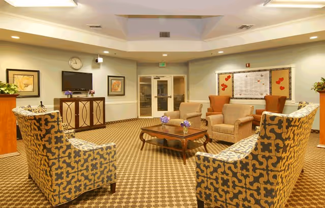 A cozy living room area in Grossmont Gardens Memory Care featuring patterned armchairs and beige upholstered chairs arranged around a wooden coffee table with small flower vases. The room has a patterned carpet, light-colored walls with framed artwork, a wall-mounted TV, a clock, and a bulletin board with red heart decorations. Double glass doors are visible at the back of the room.