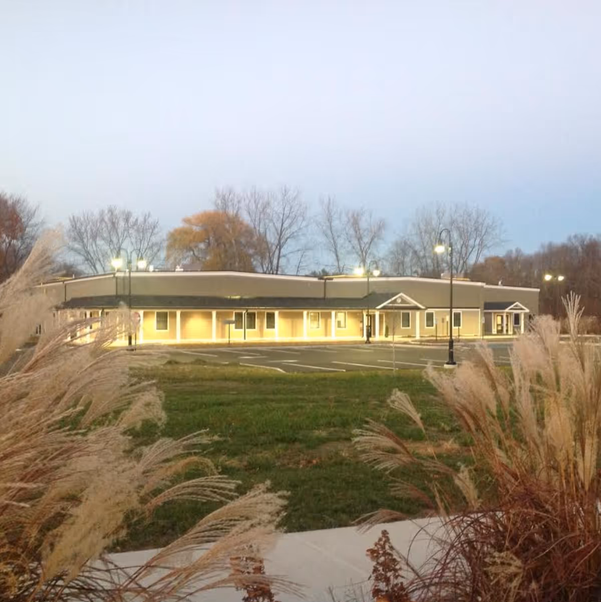 Front exterior of a single-story assisted living building lit at dusk with ornamental grasses and a parking lot in the foreground.