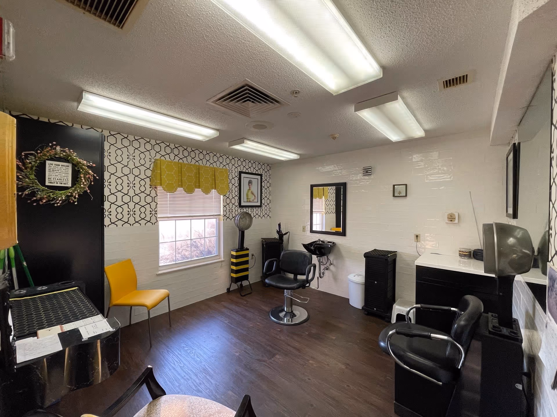 Interior of a small hair salon room with two black salon chairs, a hair washing station, a yellow chair near a window with a yellow valance, patterned wallpaper, framed pictures on the walls, and fluorescent ceiling lights.
