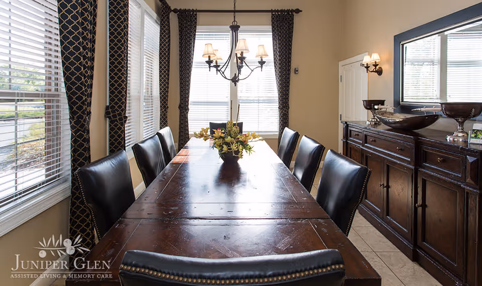 Sunlit dining room with a long wooden table, leather chairs, chandelier, sideboard and windows with blinds and curtains.