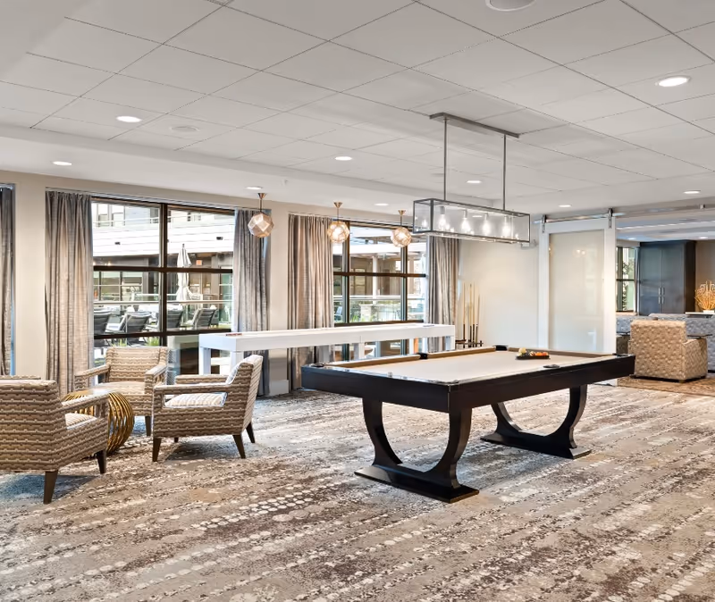 A spacious and well-lit common area featuring a pool table in the center, surrounded by comfortable armchairs and a small round table. Large windows with curtains allow natural light to fill the room. Modern pendant lights hang from the ceiling, and there is a white table along the windows. The room has a patterned carpet and a neutral color palette.