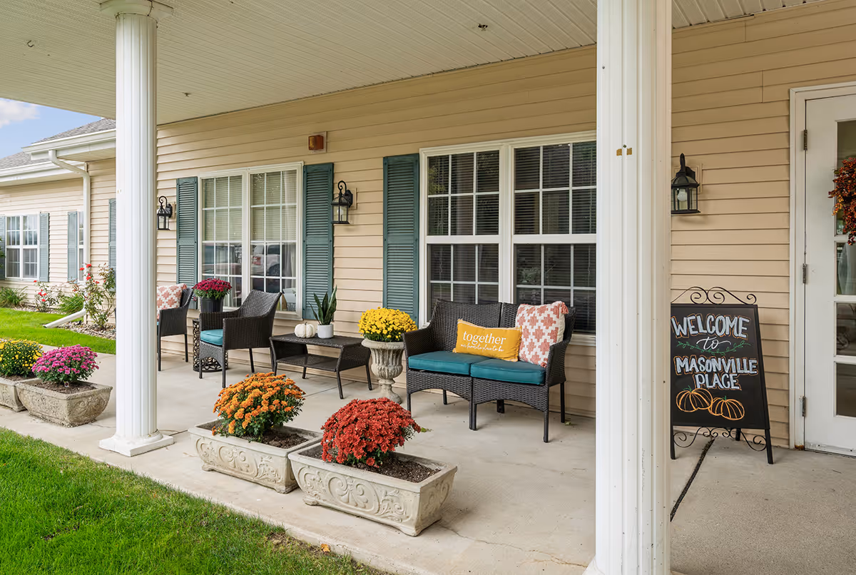 Covered outdoor patio area with wicker chairs and loveseat with cushions and pillows, decorative flower pots with colorful flowers, and a welcome sign that reads 'Welcome to Masonville Place' next to a door. The building has beige siding and blue shutters on the windows.