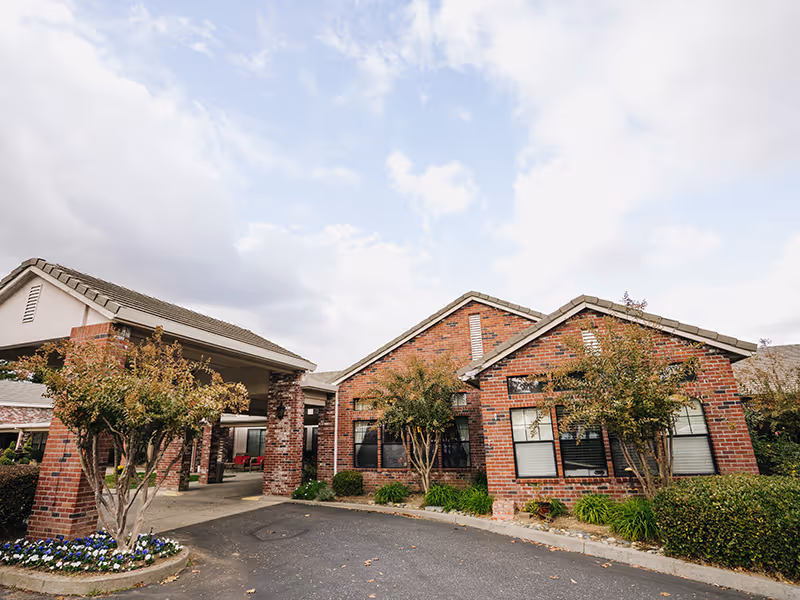 Exterior view of a brick building with a covered entrance and landscaped greenery, including trees and bushes, under a partly cloudy sky.