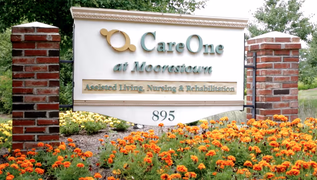 Outdoor sign for CareOne at Moorestown, an assisted living, nursing, and rehabilitation facility, surrounded by orange flowers and greenery.