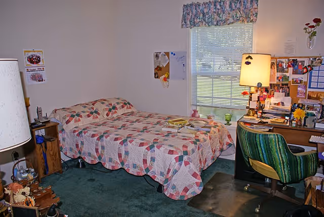 A cozy bedroom with a bed covered in a patterned quilt, a wooden nightstand with a lamp and phone, a window with floral valance letting in natural light, and a cluttered desk with a green striped chair, lamp, and various personal items and photos pinned on the wall above it.