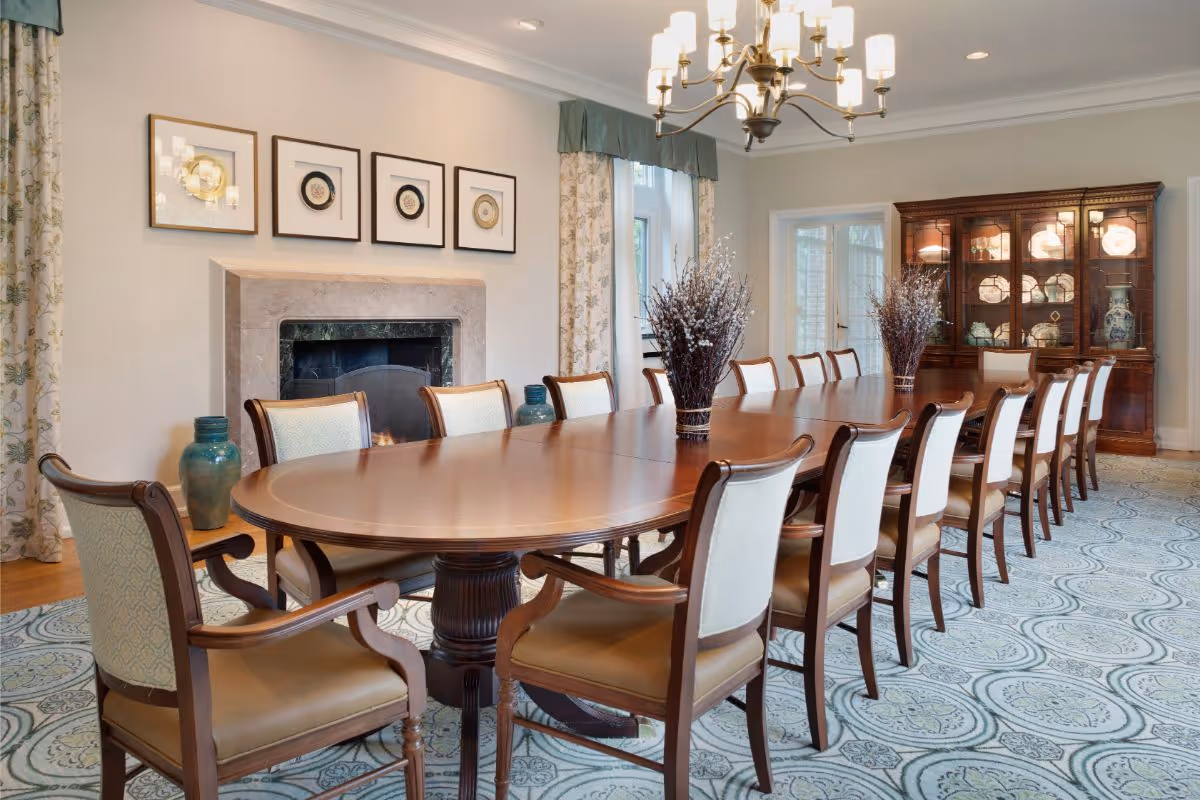 A large, elegant dining room with a long wooden table surrounded by sixteen upholstered chairs. The room features a fireplace with framed artwork above it, two large vases with decorative branches on the table, a chandelier hanging from the ceiling, patterned curtains on the windows, and a wooden china cabinet displaying plates and vases.