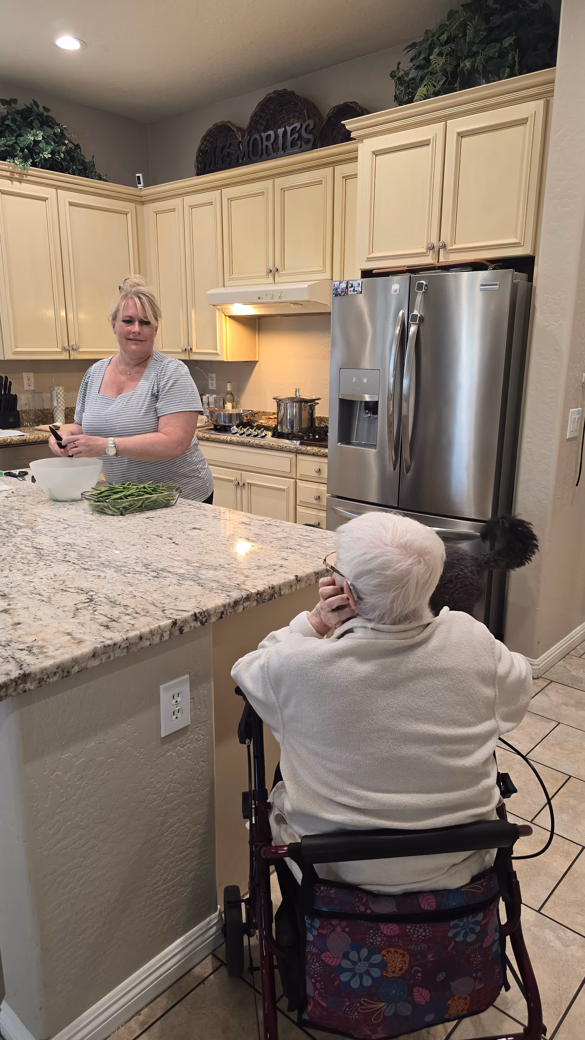 A woman prepares green beans at a kitchen island while an older adult in a wheelchair sits nearby in a residential kitchen.