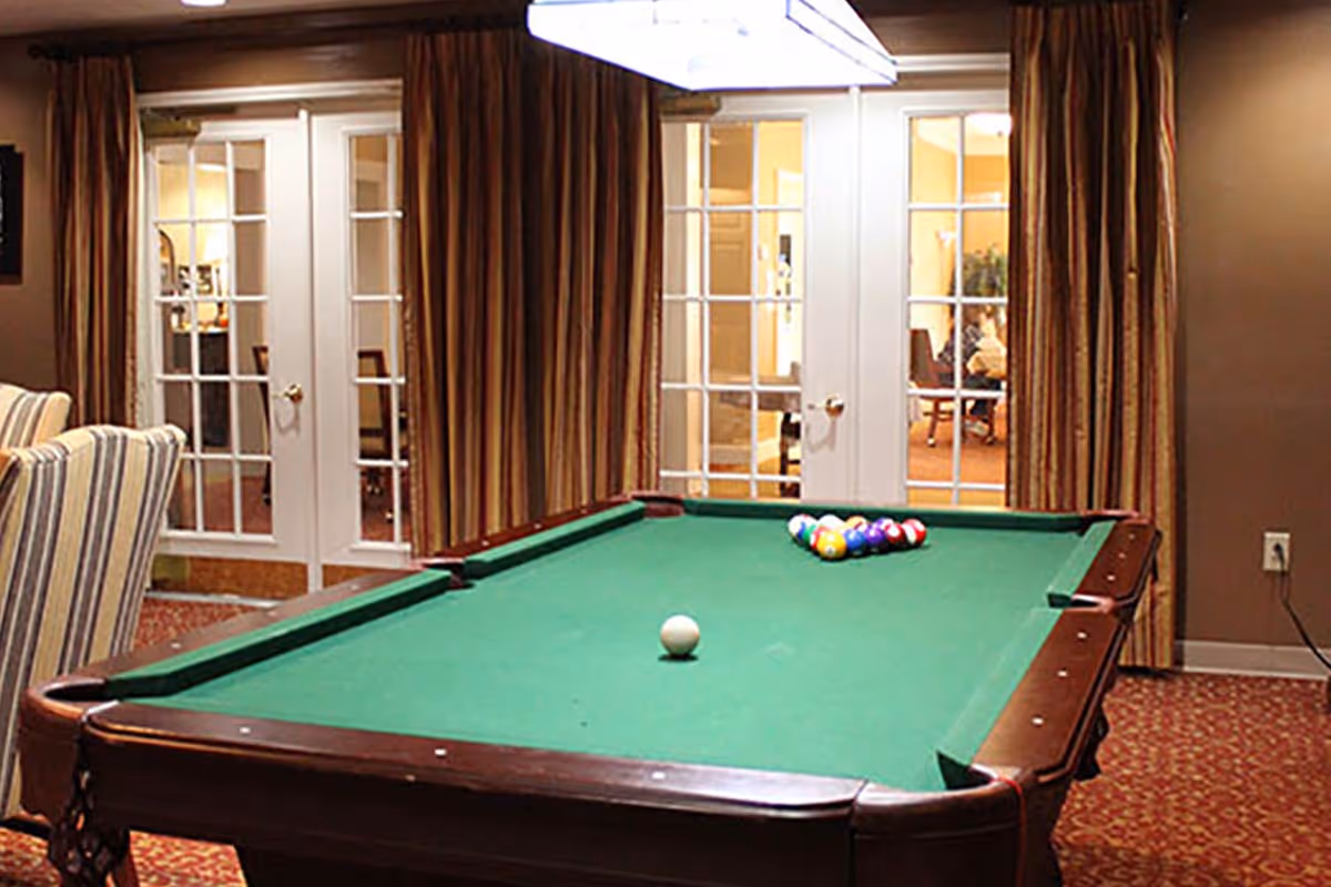A pool table with a green felt surface and a set of billiard balls arranged in a triangle at one end, located in a room with striped armchairs, patterned carpet, and glass-paneled double doors with brown curtains.