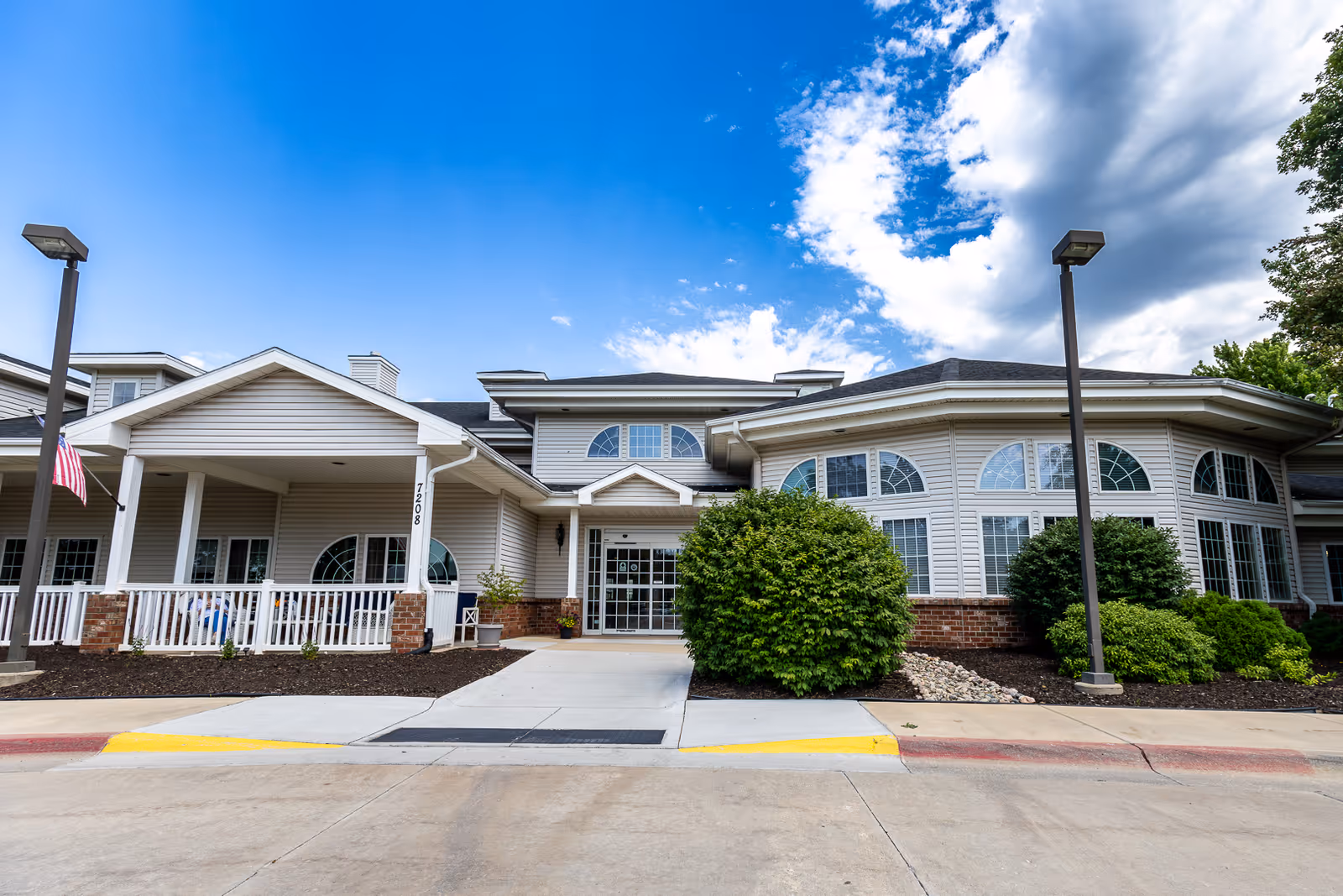 Exterior front view of Bailey Pointe Assisted Living at Van Dorn building with a covered entrance, white railing, large windows, bushes, and a clear blue sky with some clouds.