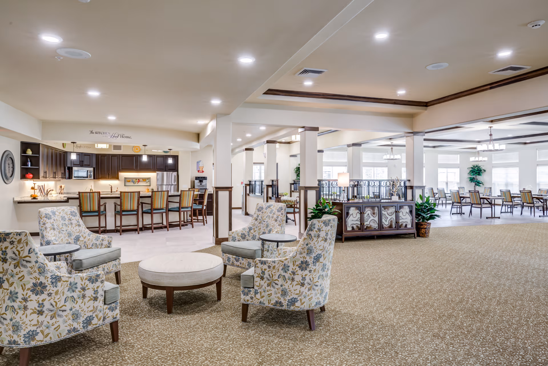 A spacious and well-lit interior of a senior living facility featuring a seating area with four floral upholstered armchairs around a round ottoman. In the background, there is a kitchen area with a counter and several bar stools, and beyond that, a large dining area with multiple tables and chairs. The space has beige carpeting, white walls, and recessed lighting.