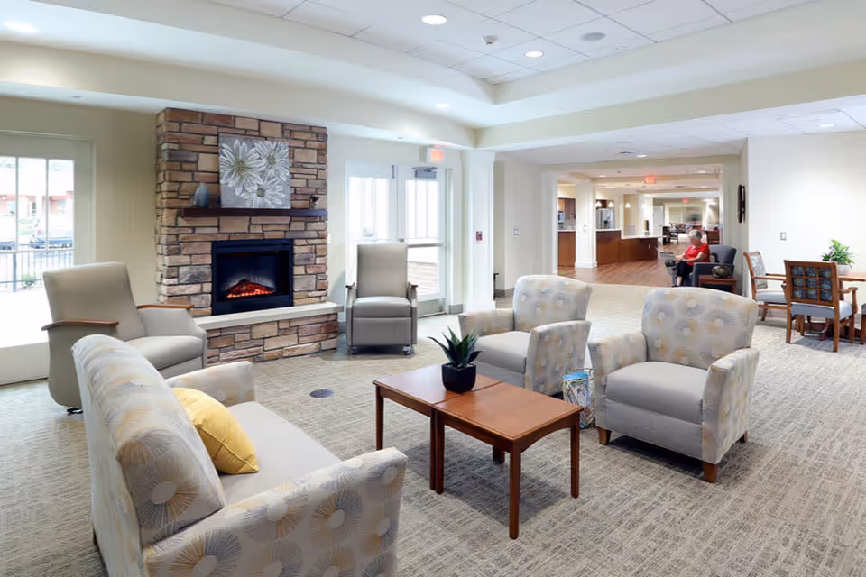 A bright and spacious senior living facility common area with comfortable armchairs and sofas arranged around a wooden coffee table. There is a stone fireplace with a floral painting above it. In the background, a person is seated on a chair near a table, and the area extends into a hallway with more seating and wooden flooring.