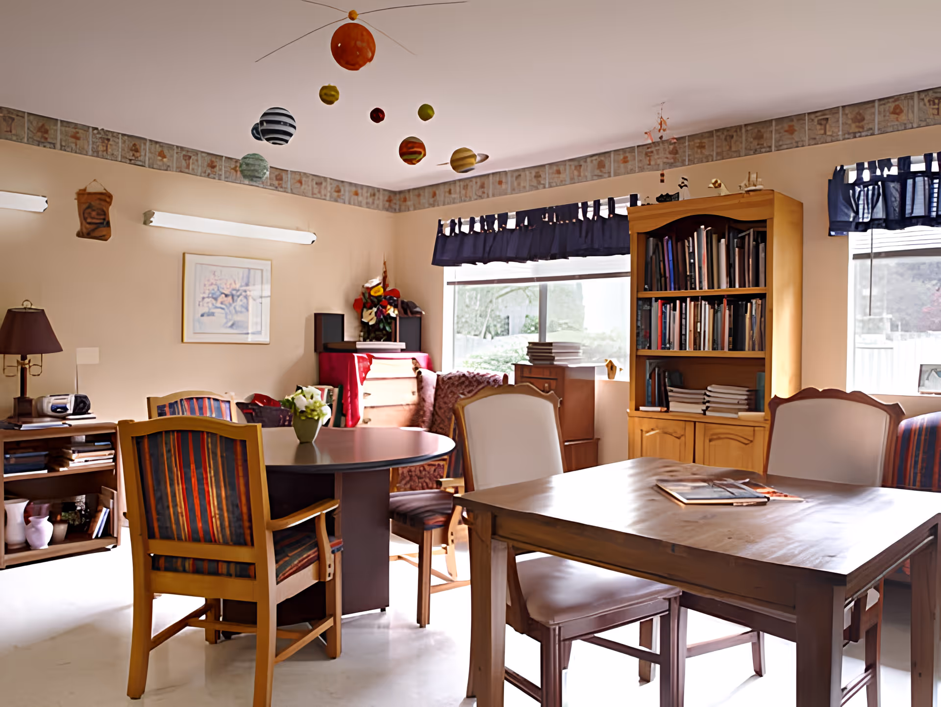 A cozy room with wooden tables and chairs, a bookshelf filled with books, a window with blue curtains, and a hanging model of the solar system from the ceiling. The room has beige walls with a decorative border near the ceiling and various decorative items including a lamp, framed artwork, and flowers.