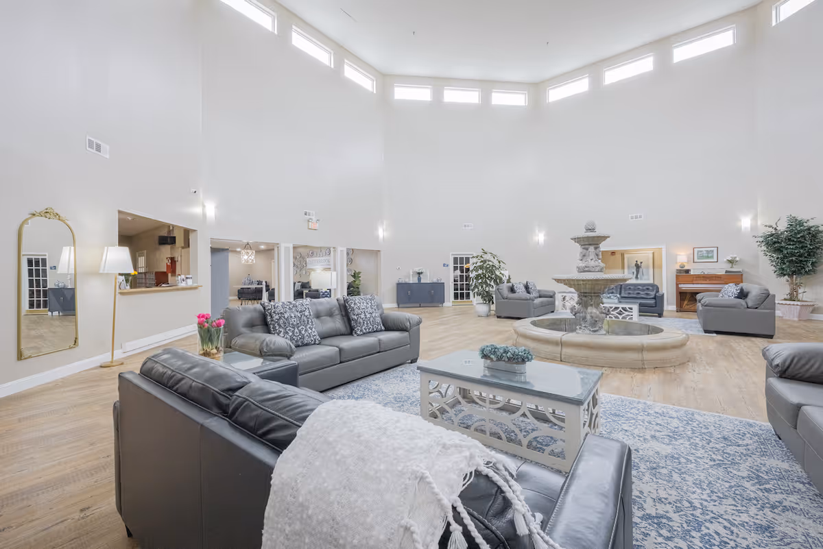A spacious senior living facility common area with high ceilings and multiple gray leather sofas arranged around a central stone fountain. The room features light wood flooring, a large patterned rug, potted plants, a floor lamp, a wall mirror, and a piano in the background. Natural light enters through high windows near the ceiling.