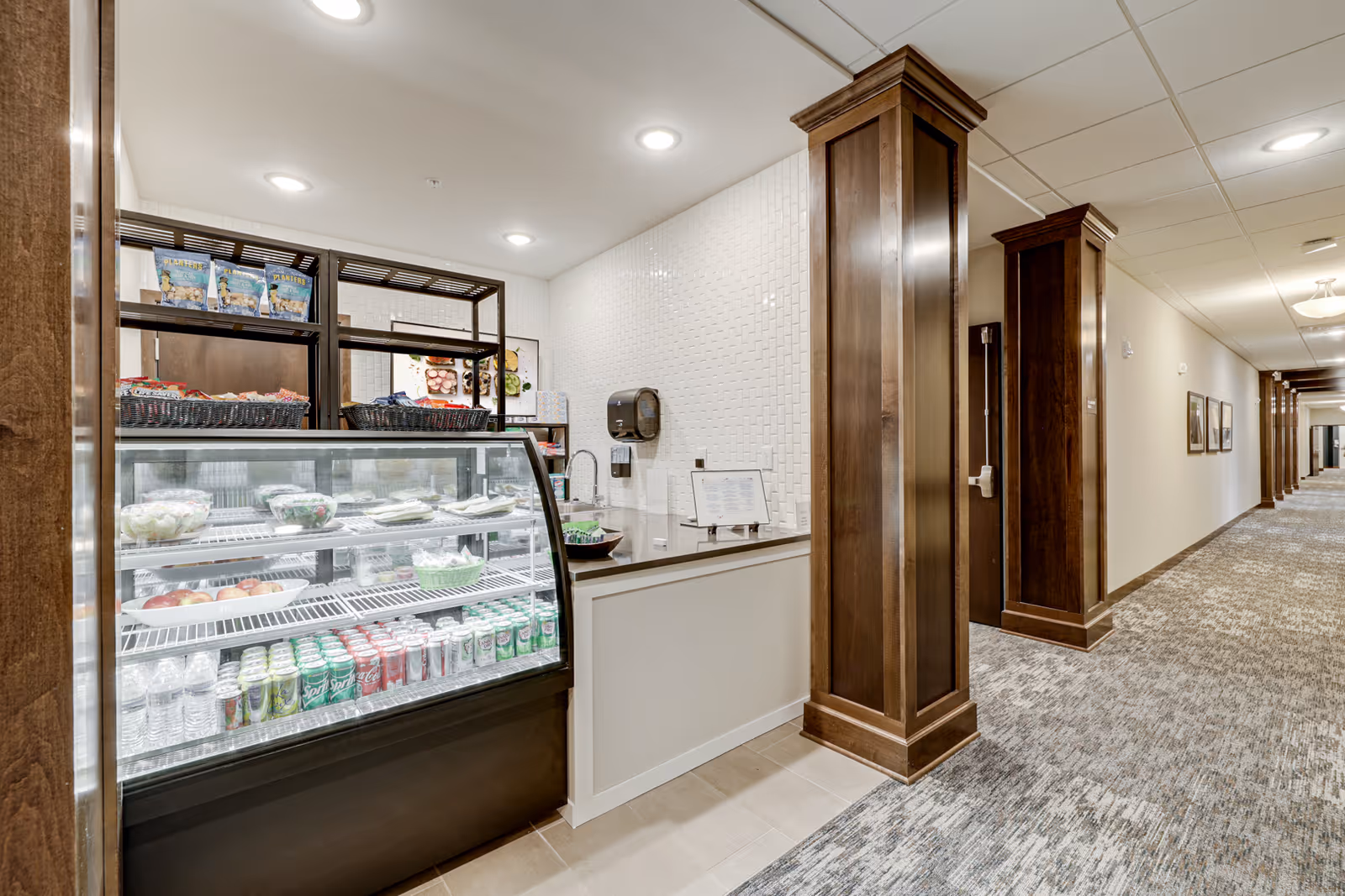 A small refreshment area in a hallway of a senior living facility with a glass display case containing bottled water, soda cans, and packaged food items. The area features white tiled walls, wooden columns, and recessed ceiling lights. The hallway extends to the right with carpeted flooring and framed pictures on the walls.