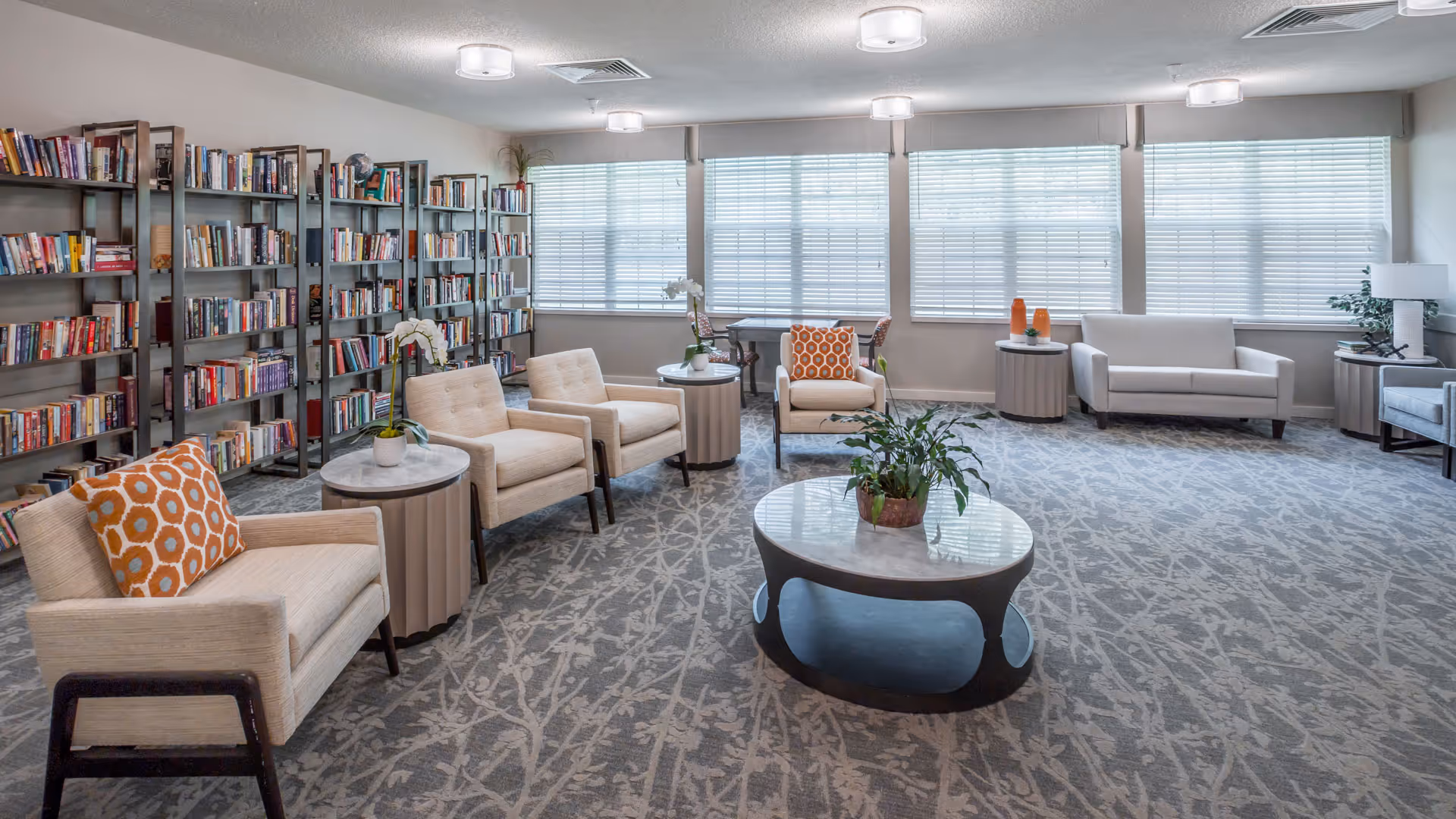 Sunlit communal library lounge with rows of bookshelves, several armchairs and sofas, side tables, and a central coffee table.
