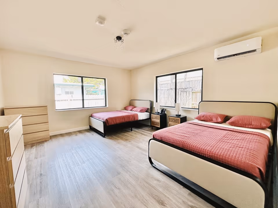 A bright bedroom with two beds, each with a red quilt and pillows. The room has two windows letting in natural light, two wooden nightstands with lamps between the beds, a wooden dresser, and light-colored wood flooring. An air conditioning unit is mounted on the wall above one of the beds.