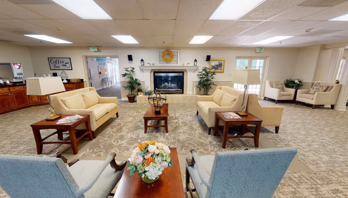 A spacious senior living facility common area with beige sofas and armchairs arranged around a central coffee table with a floral arrangement. There is a fireplace with decorative items on the mantel, potted plants on either side, and a coffee bar area to the left. The room is well-lit with ceiling lights and has carpeted flooring.