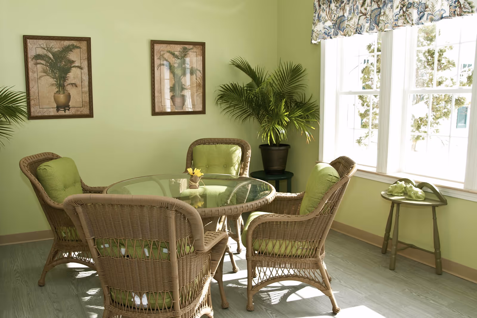 Sunlit dining nook with a round glass-top table and four wicker chairs with green cushions, potted plants, and framed botanical prints.