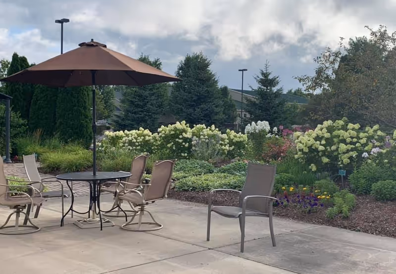 Patio with an umbrella-covered table and several chairs overlooking a landscaped garden filled with flowering shrubs and trees.