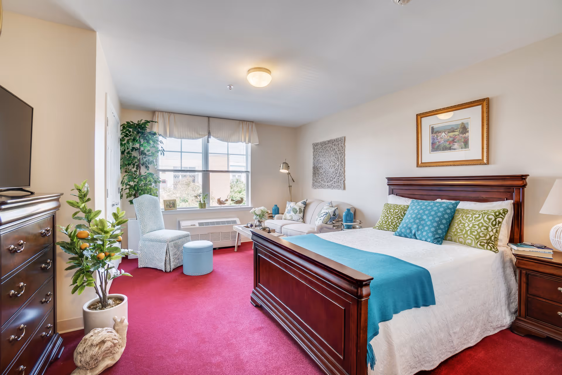 A bright and cozy bedroom in an assisted living facility featuring a large wooden bed with white bedding and colorful pillows, a wooden dresser with a TV on top, a sitting area with a white sofa, a light blue chair, and a small round ottoman. The room has a large window with white curtains, red carpet flooring, and decorative plants and artwork on the walls.