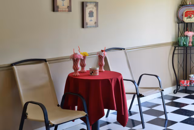 A small seating area with two beige chairs and a round table covered with a red tablecloth. On the table are three decorative milkshake-style glasses with pink and yellow flowers and a small candle holder. The floor has a black and white checkered pattern, and the wall behind has two framed pictures. To the right, there is a black metal shelf with red and green decorative items.