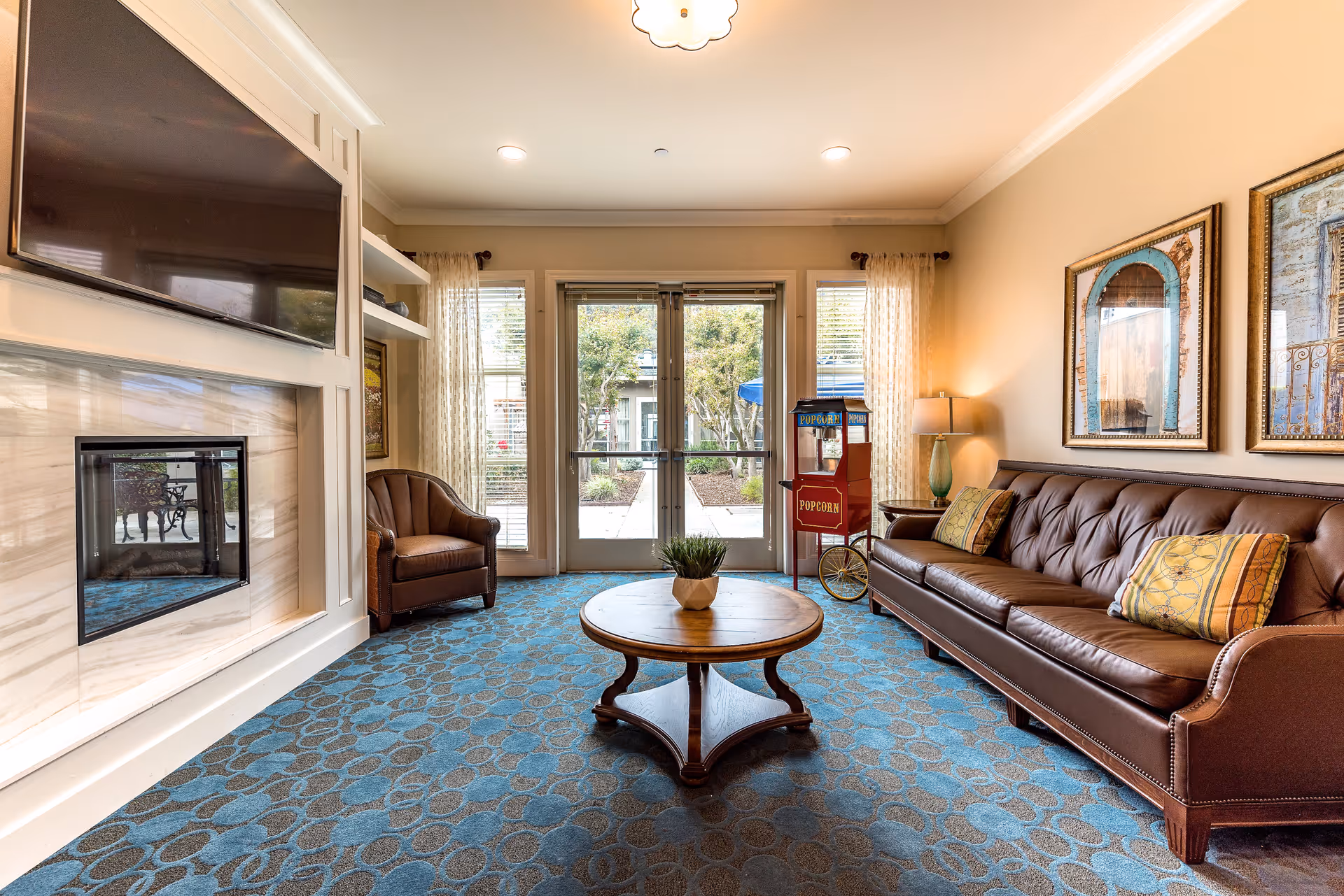 A cozy senior living facility lounge with a brown leather sofa adorned with patterned cushions, a matching armchair, a round wooden coffee table with a small plant, a popcorn machine, and a fireplace beneath a mounted flat-screen TV. Large windows and glass doors with sheer curtains allow natural light to brighten the room, and framed artwork hangs on the walls.