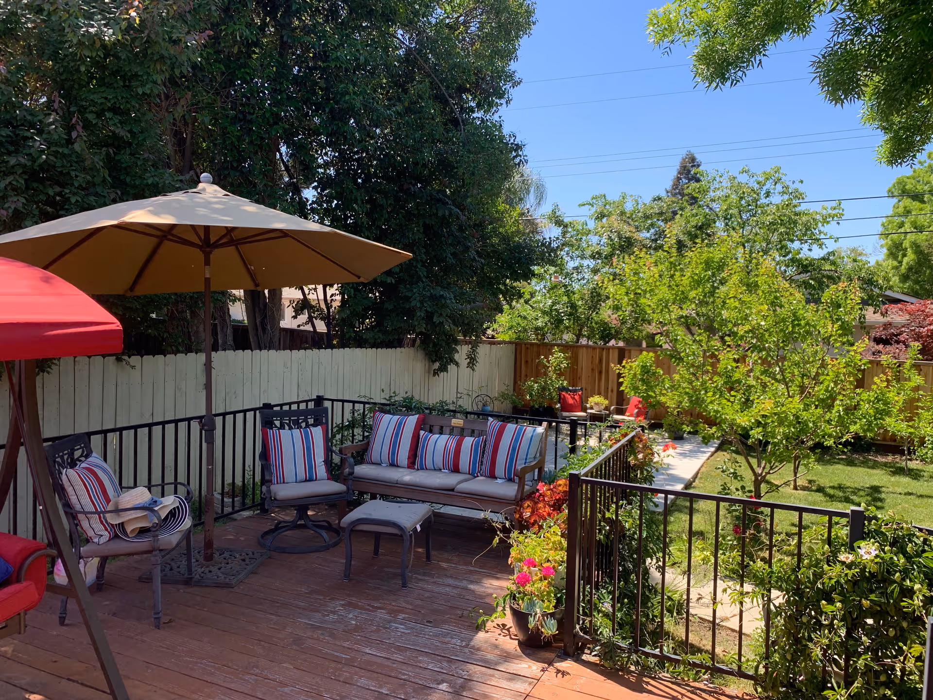 Outdoor patio area with cushioned seating including a sofa and chairs with striped pillows, a large beige umbrella, and a red canopy. The patio is surrounded by a black metal railing and overlooks a green garden with trees, plants, and a wooden fence under a clear blue sky.