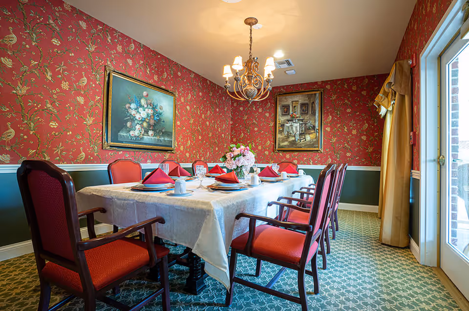 Formal dining room with a long table set for a meal, red upholstered chairs, patterned red wallpaper, chandelier, and framed paintings.