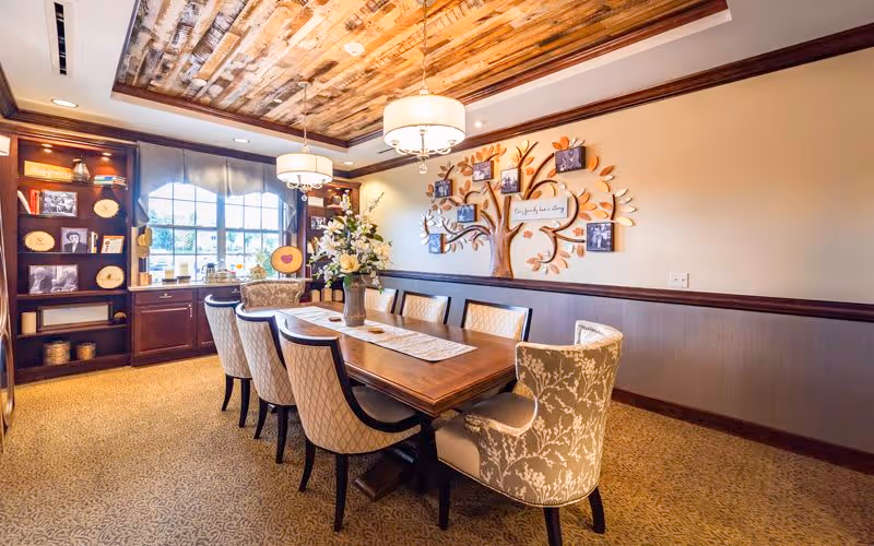 A warmly lit dining room with a long wooden table surrounded by eight upholstered chairs. The ceiling features a rustic wooden design with two hanging light fixtures. A large window with a valance allows natural light to enter. On one wall, there is a decorative tree with framed photos and a sign that reads 'Bickford Family Tree'. A built-in wooden bookshelf with various decorative items is visible near the window.
