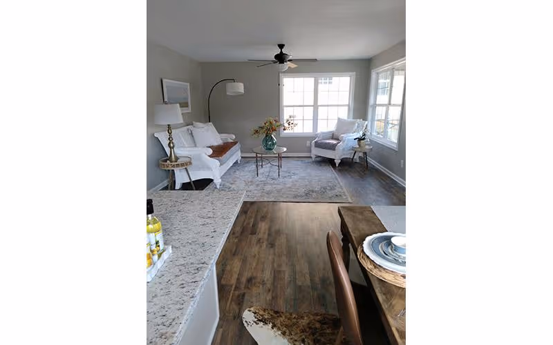 A bright living room with large windows letting in natural light. The room features a white sofa with cushions, a white armchair, a round glass coffee table with a vase of flowers, a floor lamp, and a ceiling fan. The floor is wooden with a large area rug in the seating area. In the foreground, there is a granite countertop and part of a dining table set with plates and chairs.