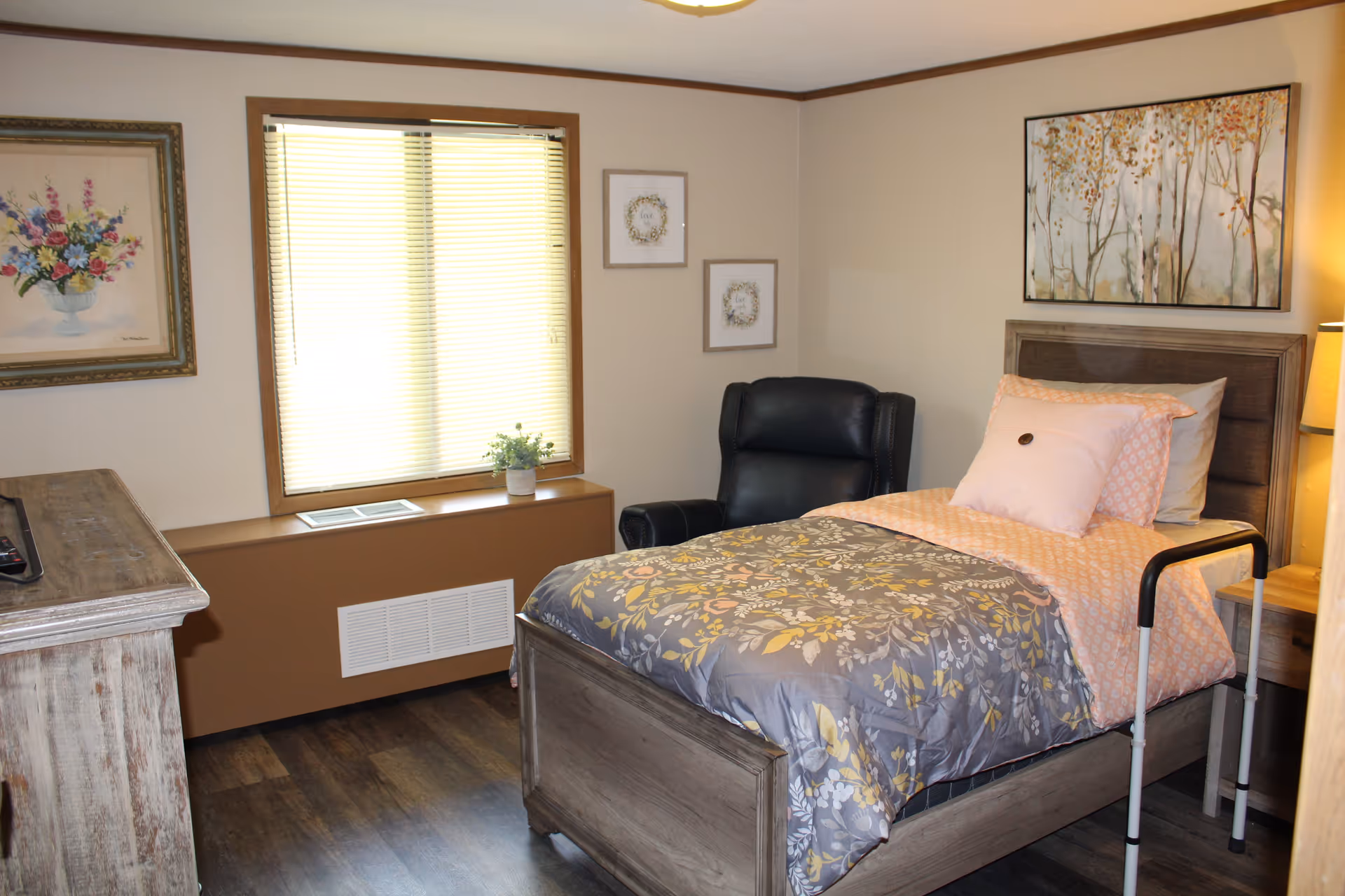 A cozy bedroom in a senior living facility featuring a single bed with floral bedding and a pink pillow, a black armchair, a wooden dresser, and a window with blinds. The walls are decorated with framed floral artwork and two small pictures. A bedside lamp is visible next to the bed.
