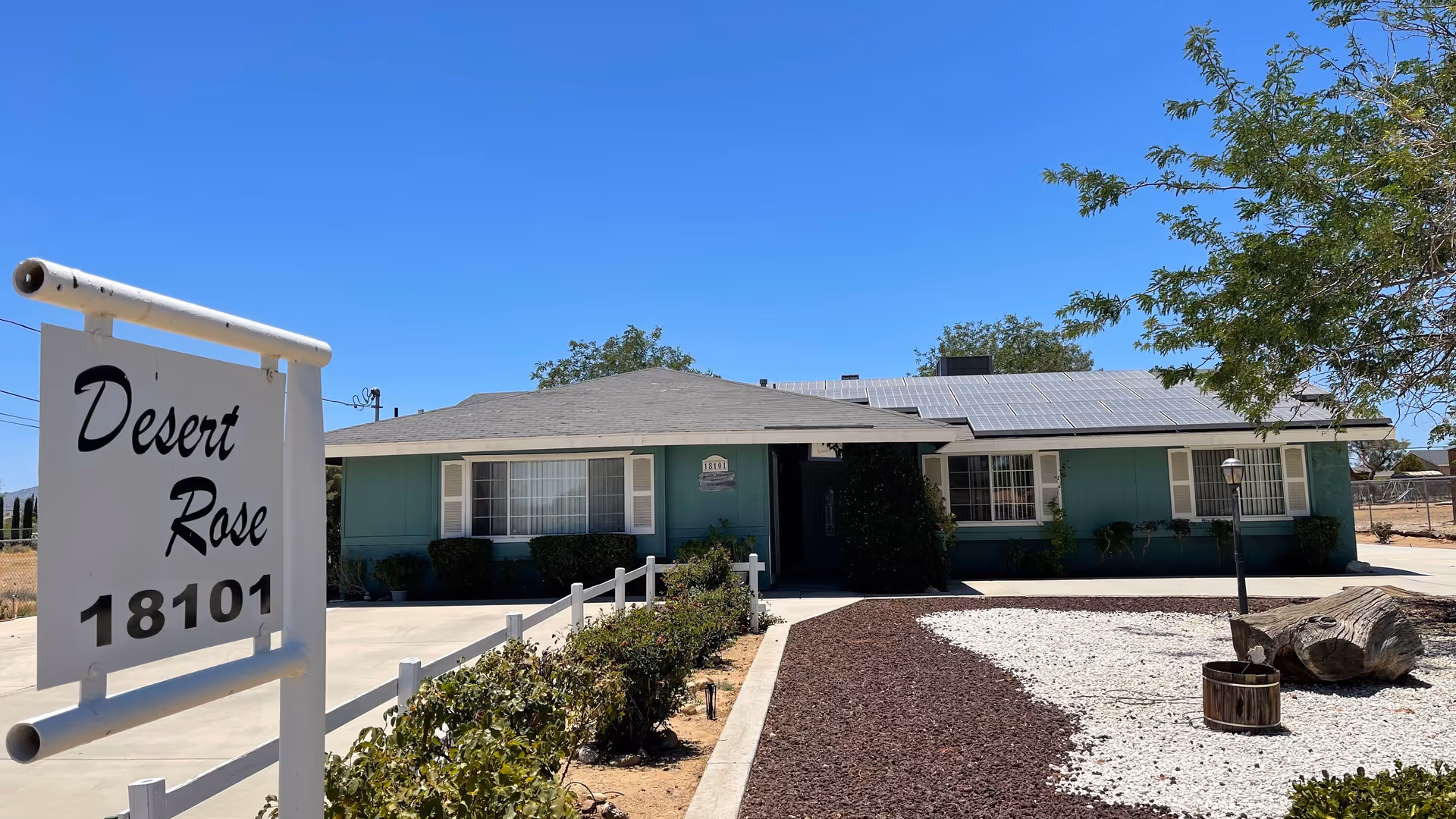 Exterior view of a single-story residential building painted teal with white trim, featuring solar panels on the roof. In front of the building is a landscaped area with bushes, a tree, and decorative rocks. A white signpost near the driveway reads 'Desert Rose 18101'.