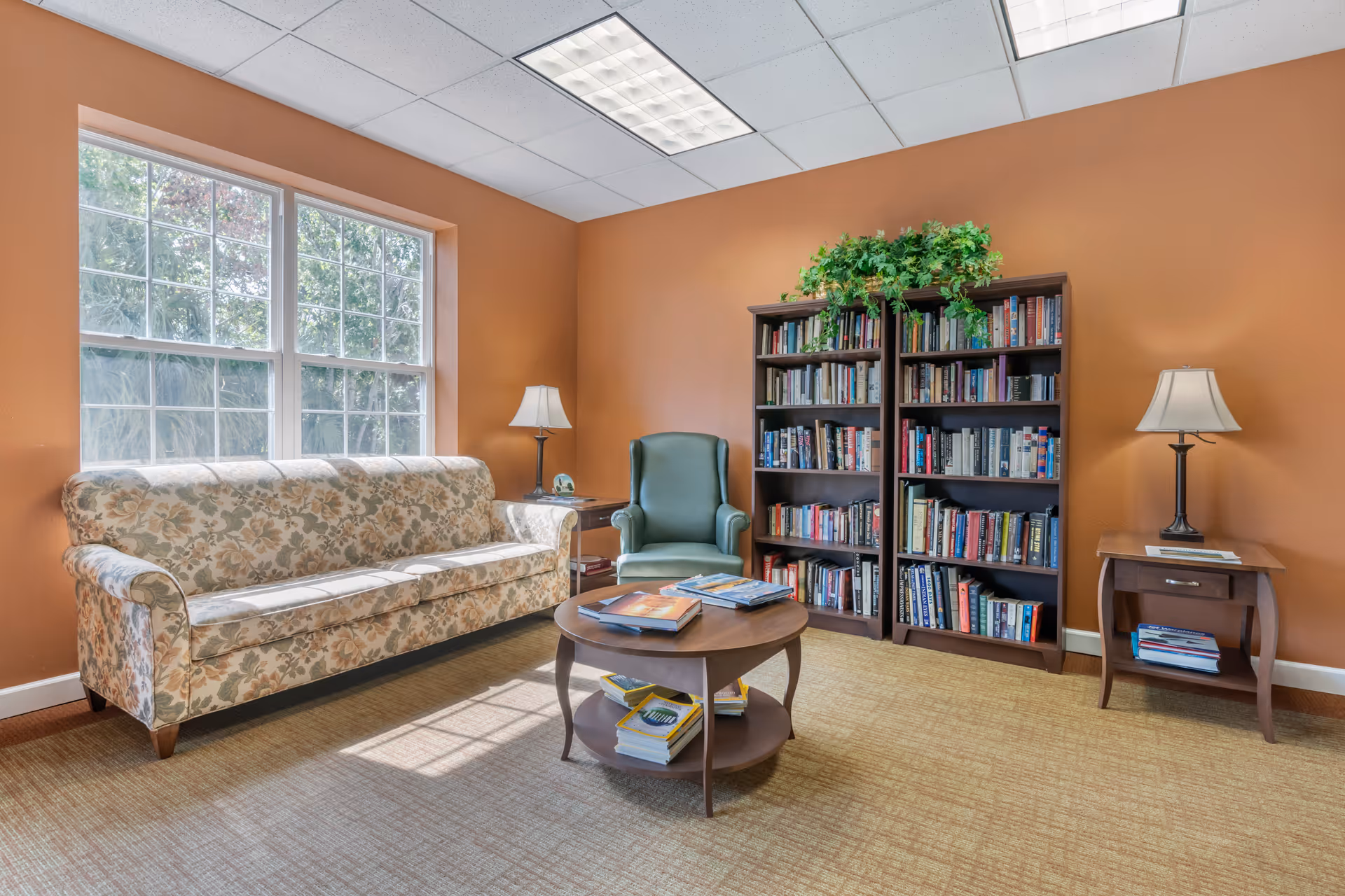 A cozy reading room with a floral patterned sofa, a green armchair, two wooden bookshelves filled with books, a round wooden coffee table with books on it, two side tables each with a lamp, and a large window letting in natural light. The walls are painted a warm orange color.