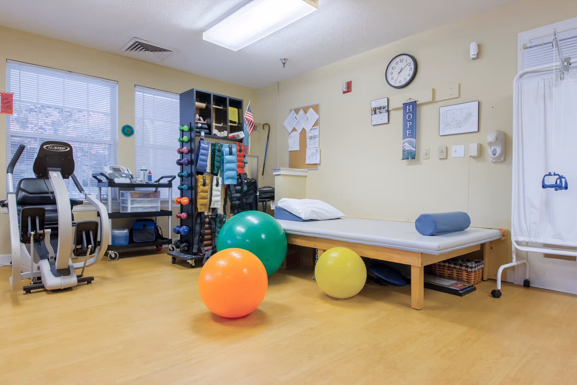 A physical therapy or exercise room with a padded treatment table, three colorful exercise balls (orange, green, yellow), a stationary exercise bike, a rack with various dumbbells and weighted belts, and a white privacy curtain. The room has light yellow walls, a clock, and a bulletin board with papers pinned on it.