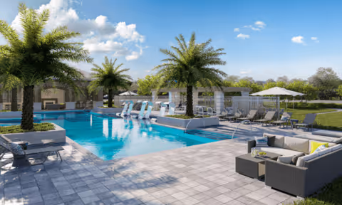 Outdoor swimming pool area with palm trees, lounge chairs, umbrellas, and a seating area with cushioned sofas under a clear blue sky.
