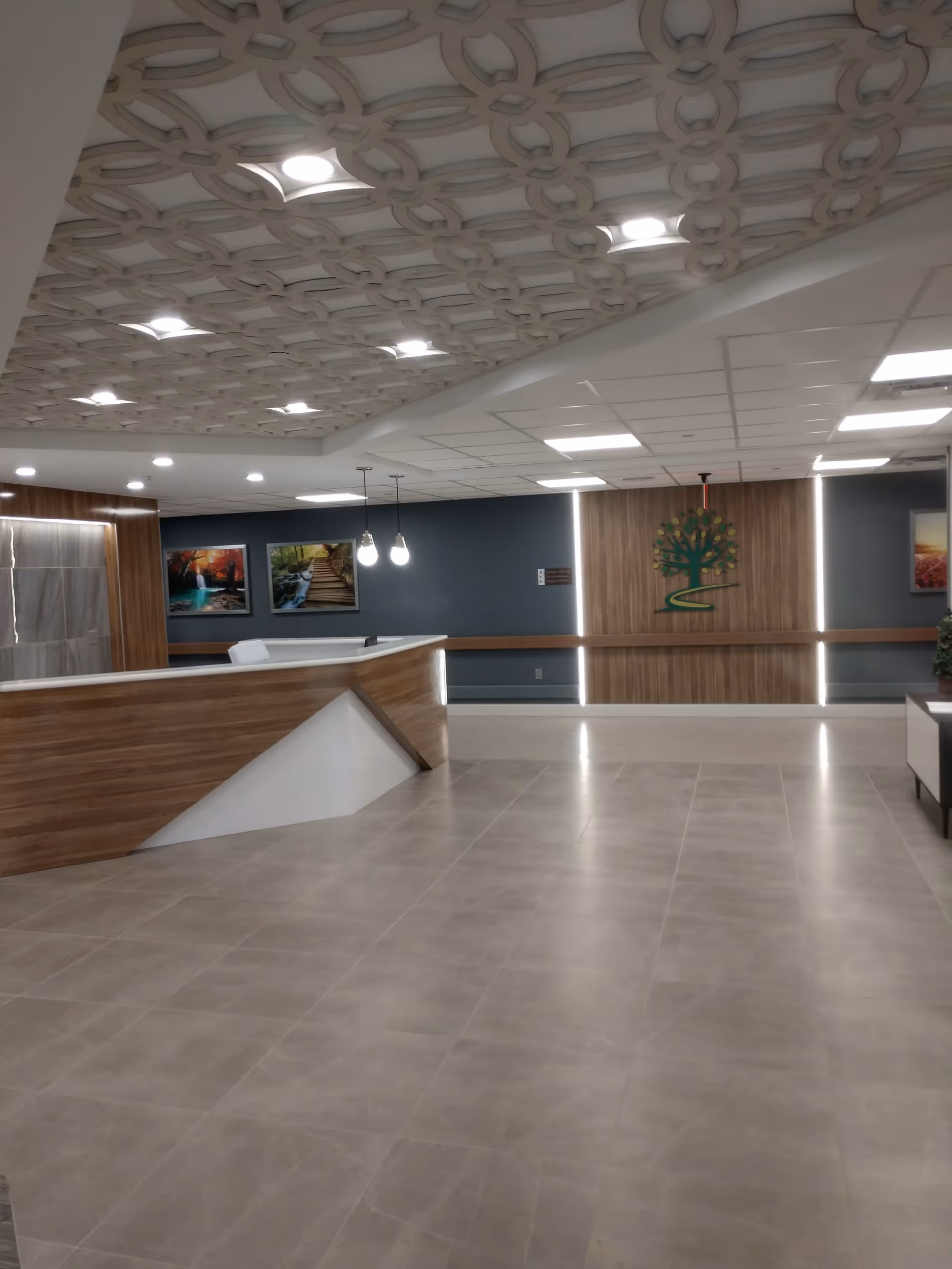 Interior view of a modern reception area with a wooden front desk, decorative ceiling with circular patterns, tiled floor, and wall art including a tree logo on a wooden panel and landscape paintings.