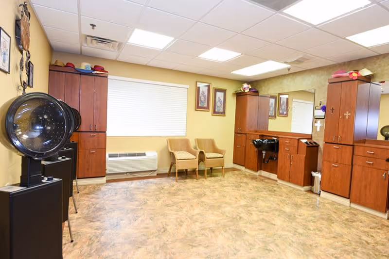 Interior of a hair salon area in a senior living facility with wooden cabinets, a large mirror, a black hair washing sink, two beige chairs, and a hair dryer chair. The room has a window with blinds, framed pictures on the walls, and a beige and brown patterned floor.