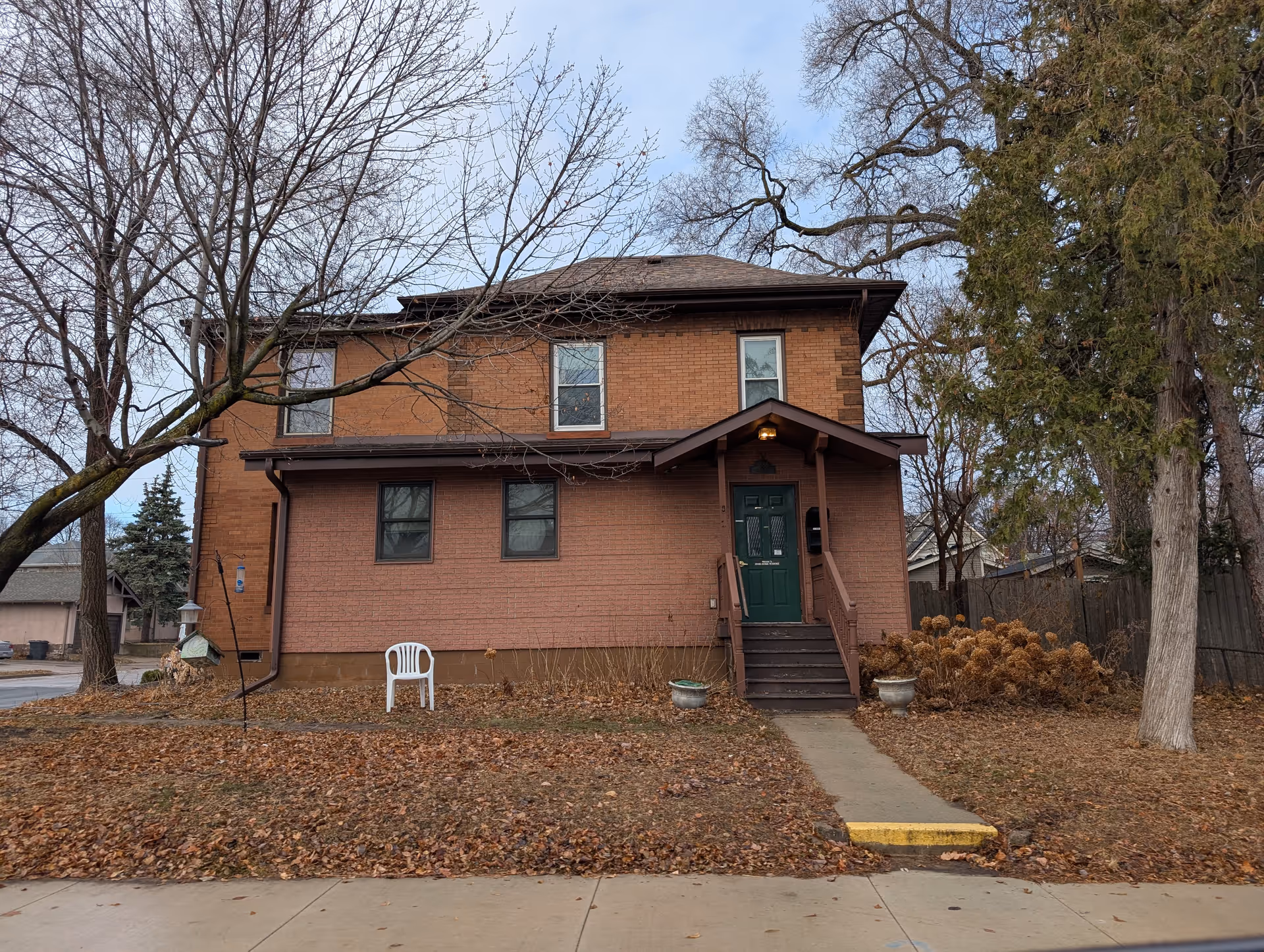 Exterior view of a two-story brick building with a green front door, a small porch with stairs, and leafless trees surrounding the property. There is a white plastic chair on the lawn and dried plants near the entrance. The sky is clear with some clouds.