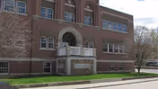 Red-brick three-story building with an arched central entrance, small balcony, and a lawn along the sidewalk.