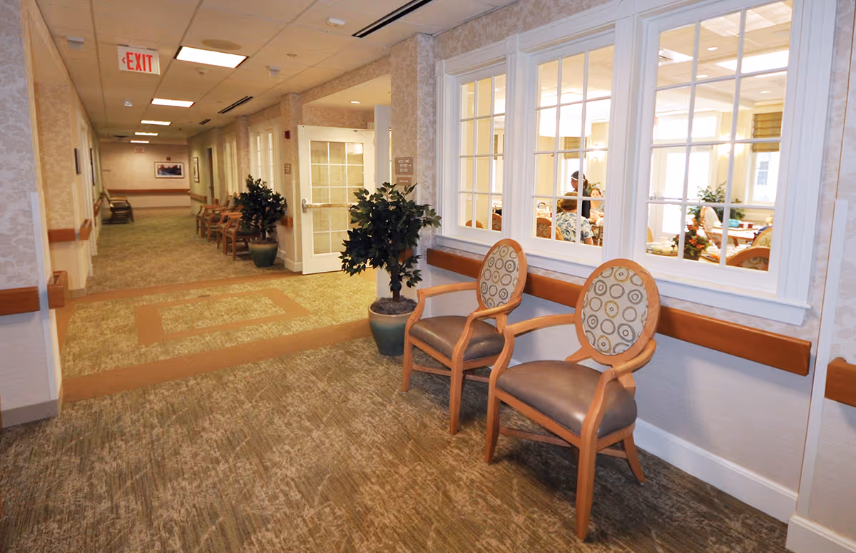 A well-lit hallway in a healthcare facility with patterned carpet and beige walls. Two wooden chairs with cushioned seats and backs are placed against the wall near large windows looking into a dining or common area where people are seated. Potted plants are positioned along the hallway, and an exit sign is visible at the end of the corridor.