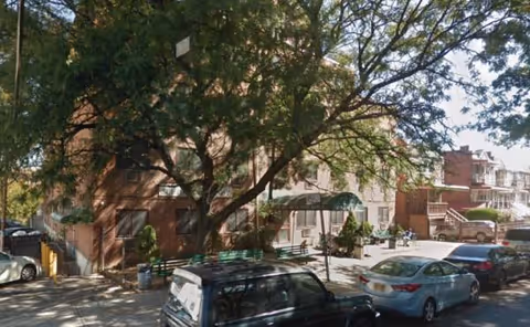 Exterior view of a brick senior living building with trees and parked cars.