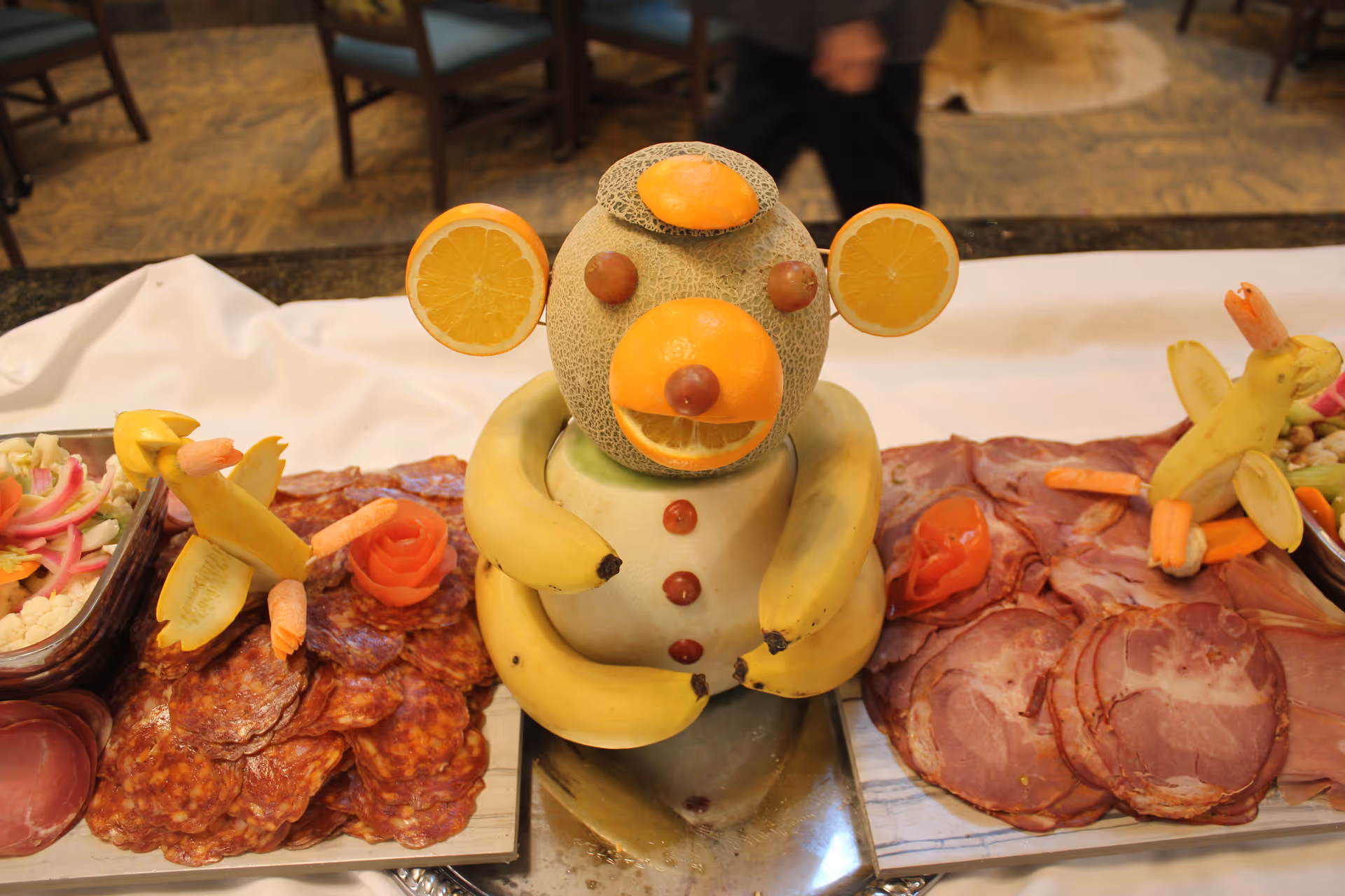 A fruit and charcuterie buffet centerpiece shaped like a smiling animal made from melons, oranges, bananas and grapes, surrounded by sliced meats and vegetables.