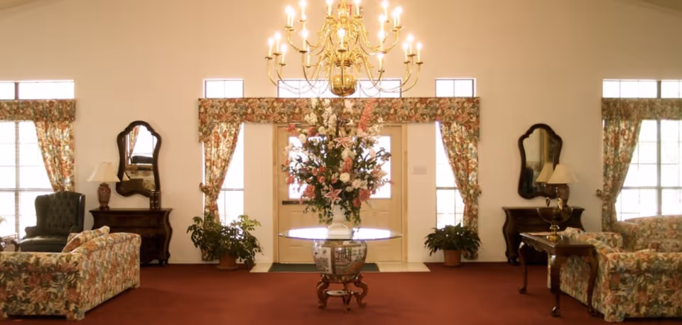 A spacious living room area with floral patterned sofas and matching curtains. A large floral arrangement sits on a round glass table in the center. Two dark wooden side tables with lamps and mirrors are positioned against the walls. The room has a red carpet and a gold chandelier hanging from the ceiling.