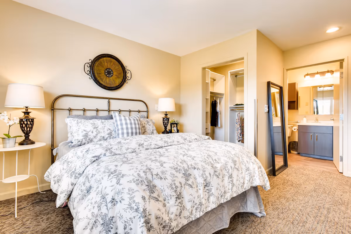 A cozy bedroom with a metal bed frame, floral patterned bedding, and two bedside lamps on white round tables. There is a decorative wall piece above the bed. To the right, there is an open closet with clothes hanging and a doorway leading to a bathroom with a sink, mirror, and lighting fixture. A full-length mirror leans against the wall near the bathroom entrance.
