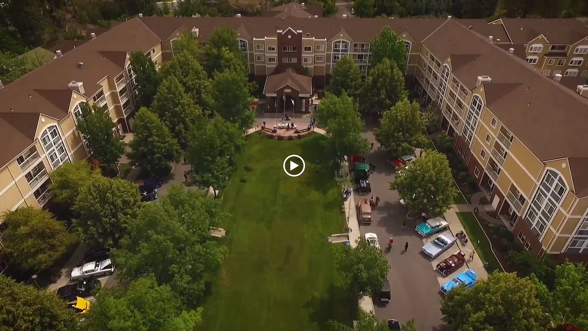 Aerial view of a U-shaped senior living building surrounding a large central lawn, driveway, and parked cars.