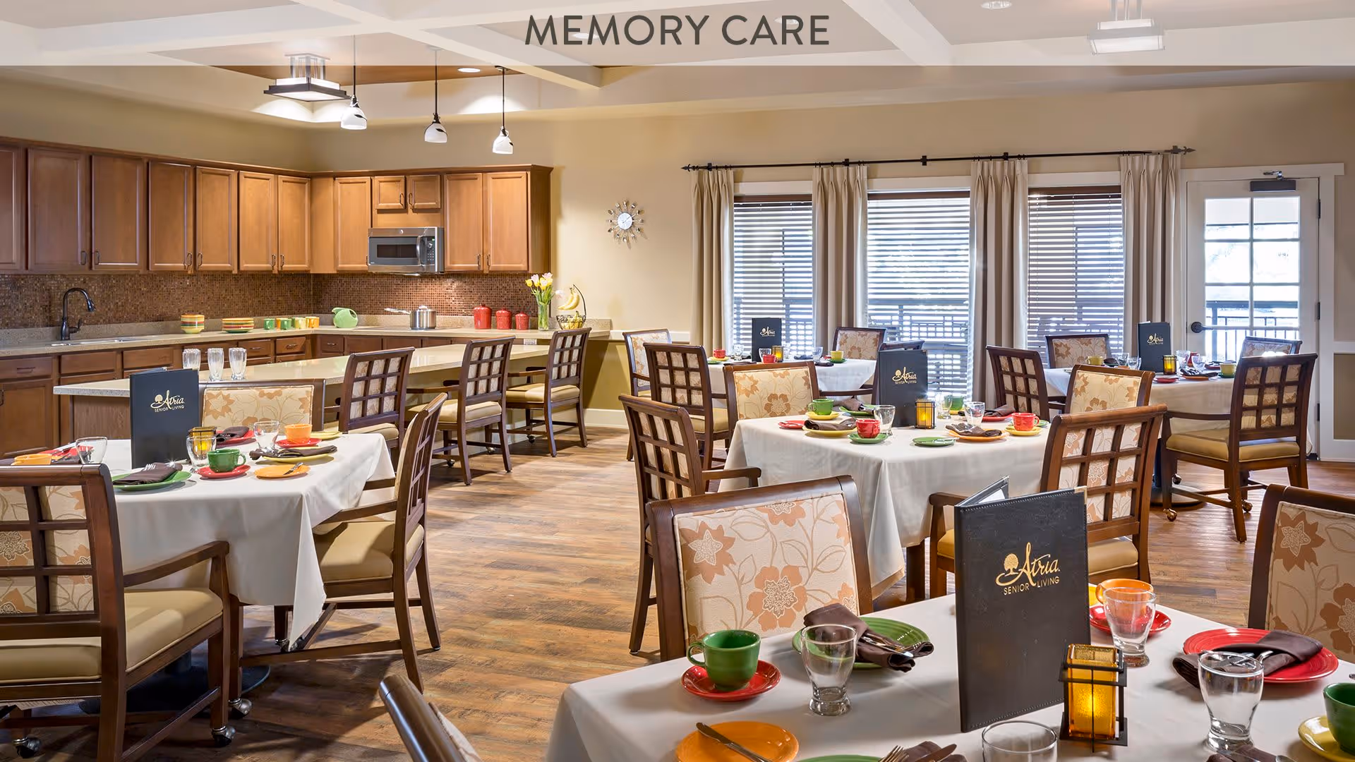 A bright and spacious dining room labeled 'Memory Care' with several tables covered in white tablecloths, each set with colorful cups, plates, and glasses. The room features wooden chairs with floral upholstery, a kitchen area with wooden cabinets and a countertop with bar stools, and large windows with blinds and curtains allowing natural light to fill the space.