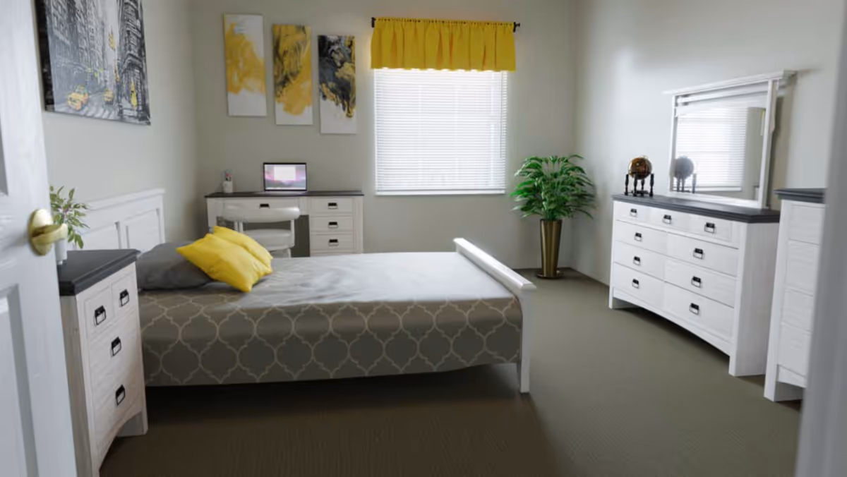 A bright and neatly arranged bedroom with a bed covered in a gray patterned bedspread and two yellow pillows. The room features white furniture including a dresser with a mirror, a chest of drawers, and a desk with a chair and a laptop on it. A window with white blinds and a yellow valance lets in natural light. There is a green potted plant in the corner and three abstract yellow and black paintings on the wall.