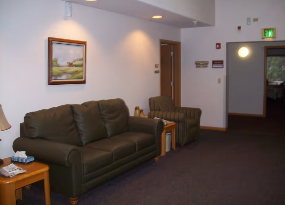A sitting area in an assisted living facility with a dark green leather couch, a striped armchair, a wooden side table with a lamp and tissue box, and a framed landscape painting on the wall. There is a hallway with doors and an exit sign in the background.