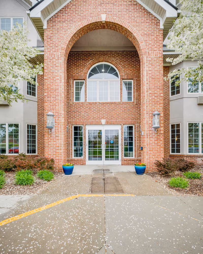 Brick main entrance with a large arched portico, double glass doors, potted plants, and flowering trees.